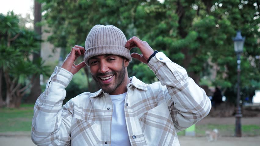 Close up portrait of young hispanic man with a beanie hat smiling and looking at camera outdoors. Front view of latin happy guy standing at park with carefree attitude. Lifestyle concept. Slow motion