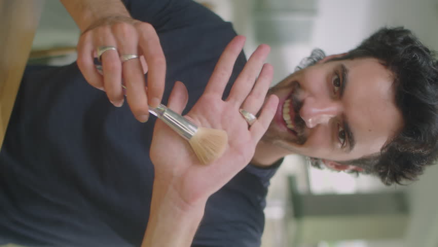 Vertical shot of male beauty blogger applying powder with cosmetic brush and explaining how to do makeup on camera while filming vlog at home