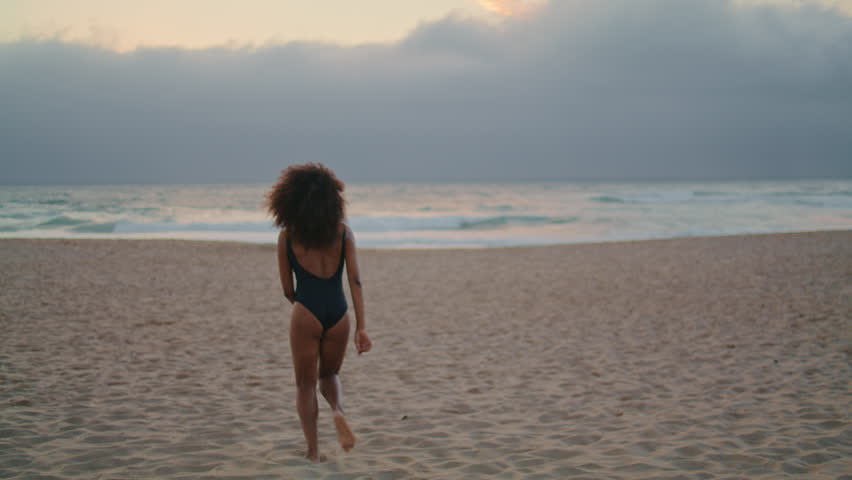 Active happy woman traveler running on sand beach raising hands up enjoying summer vacation. Curly african american girl in black swimsuit posing near ocean waves at gloomy twilight. Relax on nature. Royalty-Free Stock Footage #1100184647