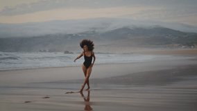 Happy active woman performer dancing on wet beach at summer twilight. Attractive slim african american girl wearing black swimsuit performing contemporary dance on seashore. Cheerful brunette have fun - Powered by Shutterstock - Get 15% off with code: PIKWIZARD15