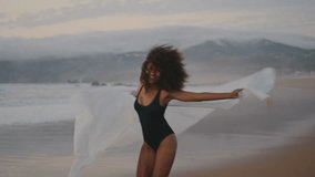 Cheerful excited girl spinning on gloomy beach with white shawl summer twilight. Curly african american woman waving pareo smiling at camera wearing black swimsuit. Happy brunette relaxing on nature. - Powered by Shutterstock - Get 15% off with code: PIKWIZARD15