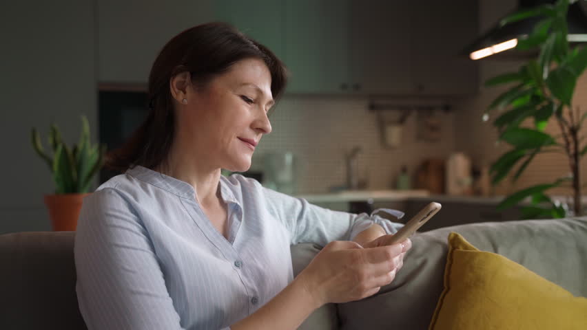 Relaxed middle aged mature woman holding smartphone looking at cellphone screen playing mobile games using social media apps, typing sms sit on couch at home. Older people and technology concept.  Royalty-Free Stock Footage #1100186341