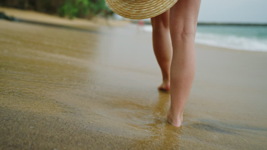 Slim female legs and feet walking along sea water waves on sandy beach. Pretty woman walks at seaside surf wearing white shirt carrying a straw hat gets splashed with waves. Girl running along shore