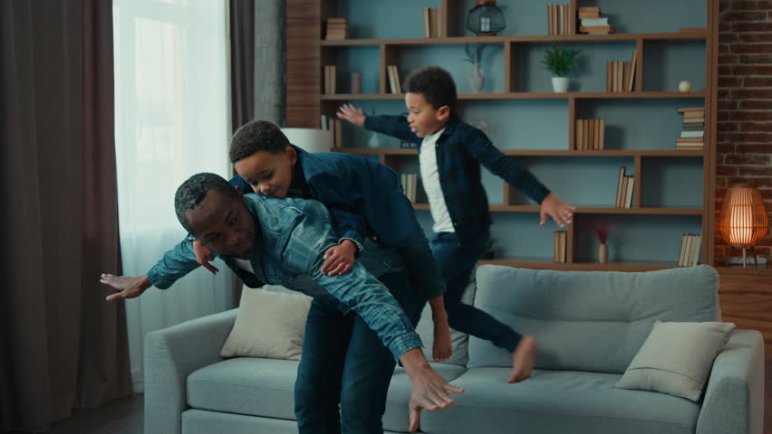 Happy African American father piggyback riding on back little preschool child son while sibling kid boy jumping on couch play airplane game. Dad with two boys children kids playing in cozy living room Royalty-Free Stock Footage #1100187021
