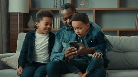 African American family adult father with two school kids children boys kids sons on sofa use funny smartphone apps amazed having fun with technology together look at phone screen video call at home