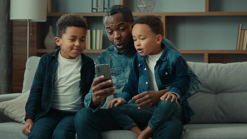 African American family adult father with two school kids children boys kids sons on sofa use funny smartphone apps amazed having fun with technology together look at phone screen video call at home Royalty-Free Stock Footage #1100187027