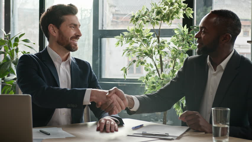 Two successful diverse businessmen sit at office desk with laptop happy shake hands. Caucasian man salesman manager handshake satisfied African American client customer investor at business meeting Royalty-Free Stock Footage #1100187039