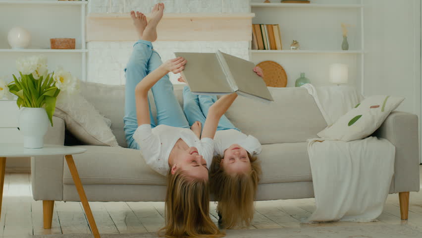 Cheerful Caucasian mother lying upside down on comfortable sofa with little child girl kid daughter reading book read together looking at pictures in textbook album family enjoy weekend at home couch Royalty-Free Stock Footage #1100187101