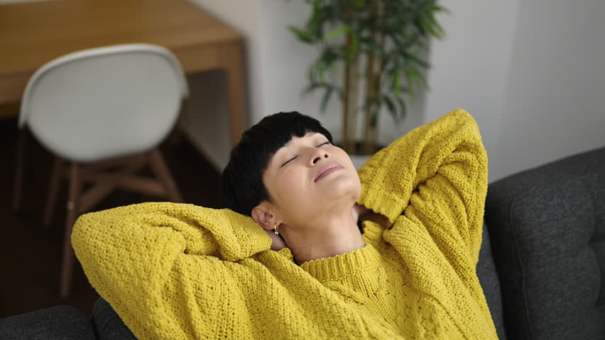 Young chinese woman relaxed with hands on head sitting on sofa at home