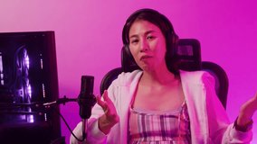 Young Asian female gamer sit on gaming chair wear headset use looking at camera online live stream talk with audience review upcoming new game release sharing to follower on computer at house.