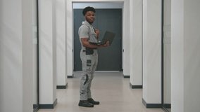 African-American security cameras installer in uniform stands in corridor with laptop, looks at camera. He sets up CCTV cameras in office using computer software. Surveillance system concept. Vertigo