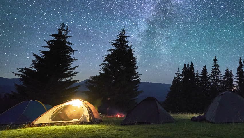 Night camping with five tourists illuminated tents. Hiker having a rest near campfire and forest, cooking dinner on fire. Timelapse of the starry sky with the Milky Way. Concept of tourism and nature.