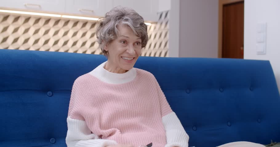 Senior woman watches something exciting on TV and makes a call by the phone, 4k