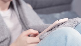 Teenager girl typing on phone in pink case. Relaxed girl in grey comfortable blanket using smartphone for texting and social media. Tap to click on screen