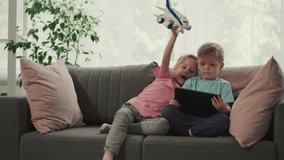 Sister bothers with airplane toy younger brother while watching videos on tablet. Cute children use modern gadget sitting on sofa in living room