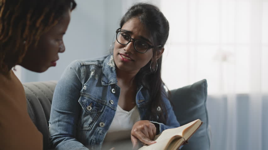 International lady students looks for information for joint educational project. African American and Indian women check book sitting on sofa in room closeup | Shutterstock HD Video #1100210315