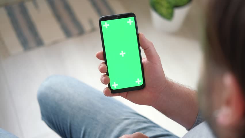 Young man sitting at home holding smartphone green mock-up screen in hand. Male person using chroma key mobile phone. Vertical mode. Touching, swiping display, tapping, surfing Internet social media | Shutterstock HD Video #1100210981