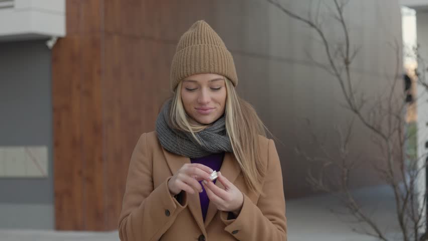 Positive young female in warm coat smiling and looking away while smearing lip balm on lips during stroll on city street