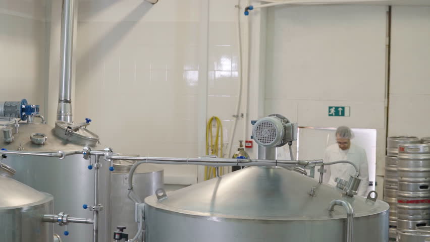 Worker is coming up to the tank brewing the barley grain. Worker is opening the lid of the tank during the brewing process. Worker is stirring the brewing grain inside of the processing tank.