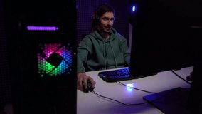 Zoom in real time of young concentrated man in headphones speaking during video game on computer in dark room illuminated by neon lights