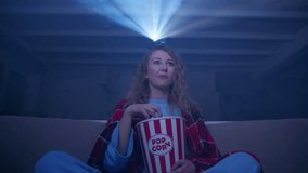 Woman with curly hair watching a concert on a projector, eating popcorn and dancing on the couch at home. High quality 4k footage