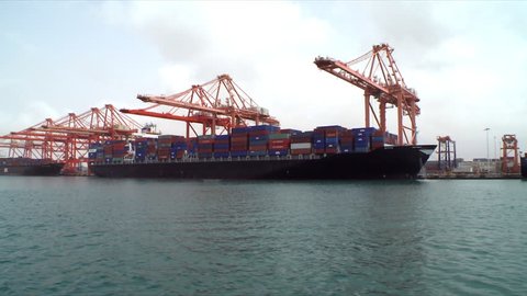 A tracking shot past container ships docked at a port in Oman with a row of cranes behind. 