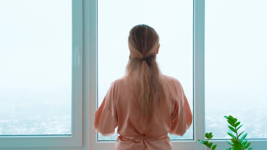 Mysterious dark silhouette of woman goes to balcony panoramic window, enjoys looking view from outside. Girl back view steps, touches glass with hands without face. Winter gray sky snow is falling. 4k Royalty-Free Stock Footage #1100222689