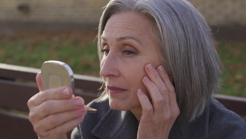 Senior gray-haired woman in elegant coat looking at her wrinkles in pocket mirror, contemplating about lifting and botox injections, anti-aging care for skin, rejuvenation