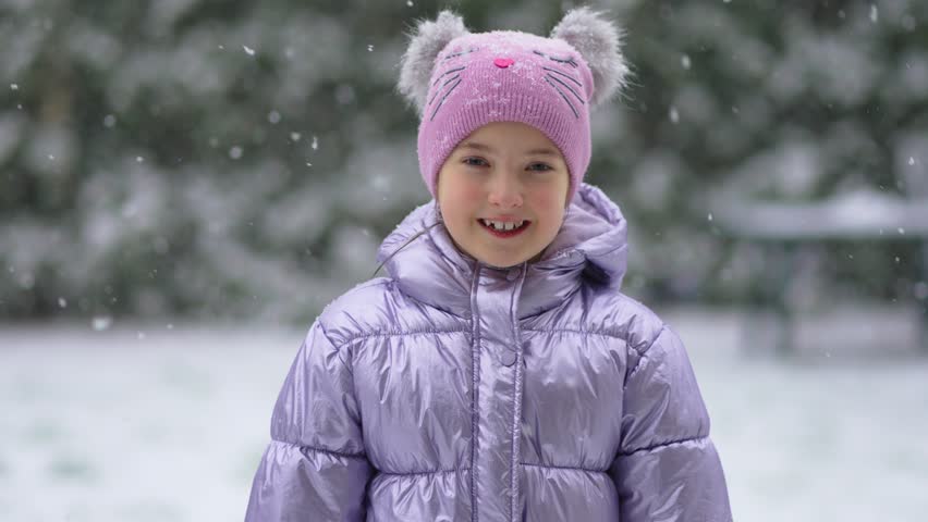 Medium close-up portrait of cheerful girl in warm clothes standing against pine wood on snowy winter day smiling at camera slow motion. Snowstorm and blizzard in winter season