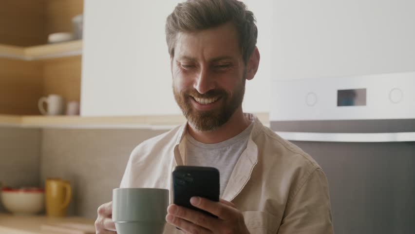 Unemployed disappointed bearded caucasian man with coffee standing at kitchen interior getting bad news on phone call and going away.  Royalty-Free Stock Footage #1100237809