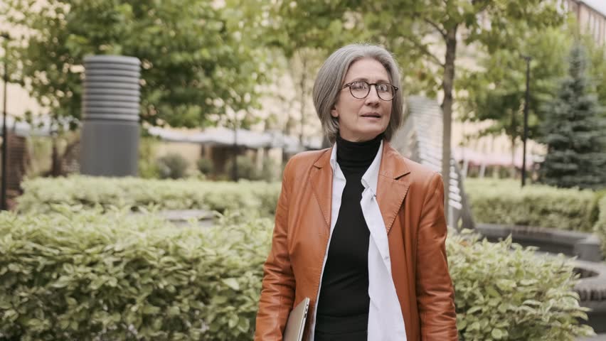happy elderly business gray-haired woman 60s years old in glasses walking down the street and smiling looking at the camera Royalty-Free Stock Footage #1100238559