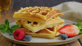 Sprinkling powdered sugar on soft Belgian waffles served on plate, pile of fluffy waffles garnished with fresh berries place on wooden table, close up slow motion video clip, 4k footage