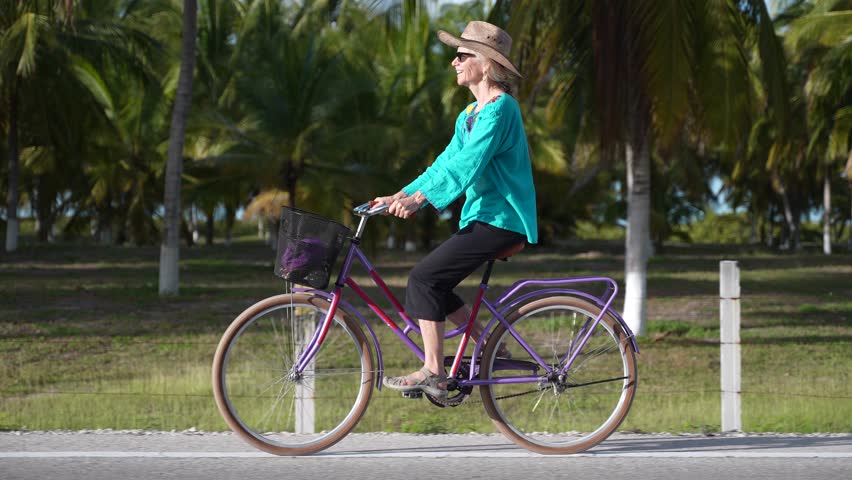 Slow motion side view portrait of pretty senior mature woman smiling wearing sunglasses, a hat, and ethnic clothes on a bicycle at sunset holding out her arm in happiness.