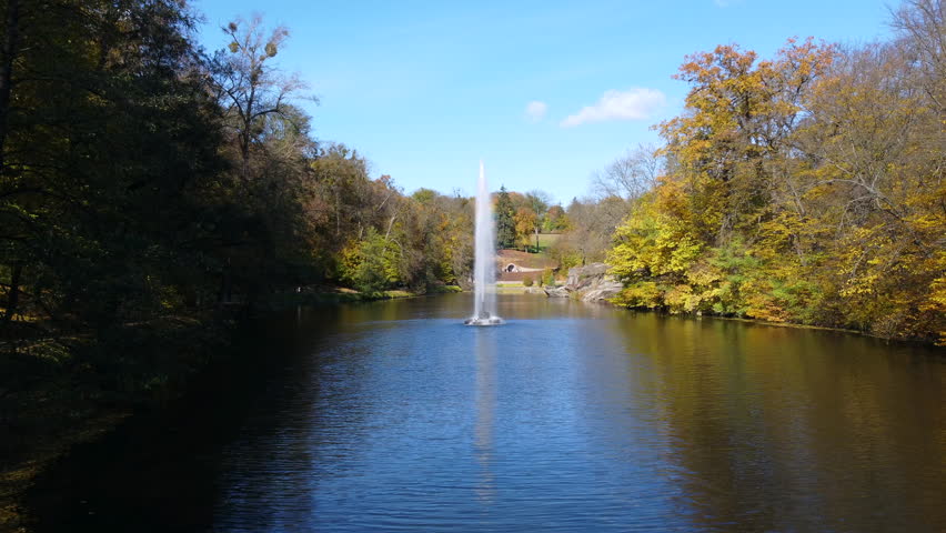 Fountain with rainbow in middle of lake between trees with yellow leaves in park on sunny autumn day. Decorative fountain in center of lake. Natural park arboretum in autumn. Natural landscape. Aerial