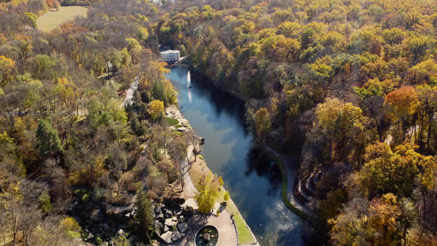 Beautiful panoramic autumn landscape park, many trees with yellow leaves, lake with fountain in center, architecture, big stones, paths walkways, people walking on sunny autumn day. aerial drone view