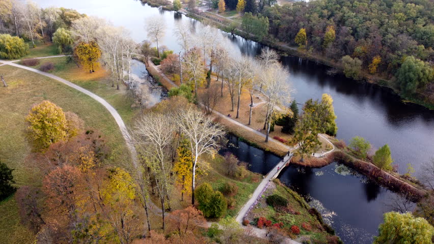 Flying over autumn park. Many trees with yellow green and fallen leaves, lakes, river, people walking along dirt paths in park on autumn day. Top view. Aerial drone view. Beautiful natural background