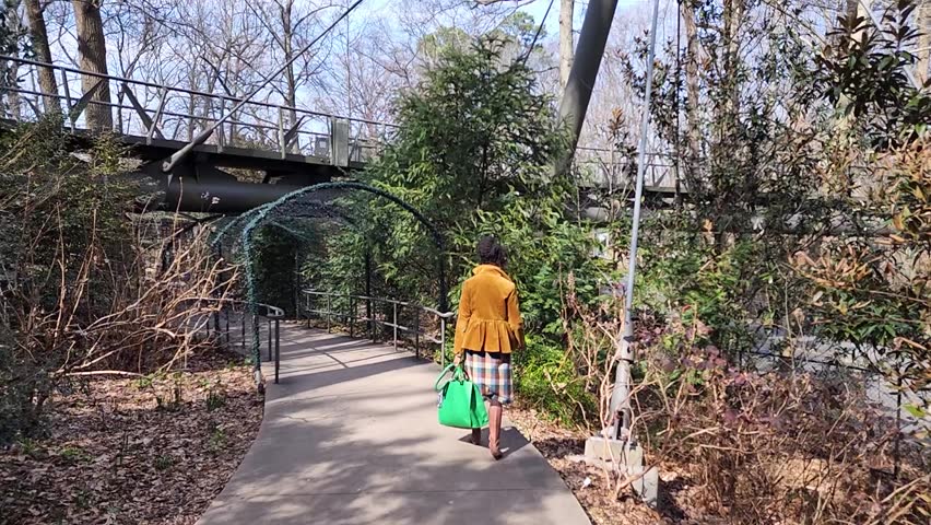 Slow motion footage of an African American woman with long sisterlocks wearing a brown coat and sunglasses walking in the garden with trees and plants at Atlanta Botanical Garden in Atlanta Georgia