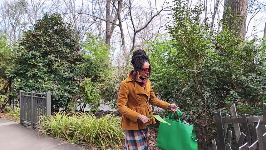 Slow motion footage of an African American woman with long sisterlocks wearing a brown coat and sunglasses walking in the garden with trees and plants at Atlanta Botanical Garden in Atlanta Georgia