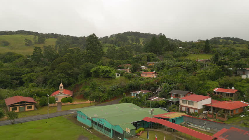 Backwards ascending reveal of sports area in small town. Football pitch and surrounding houses, green landscape in background. La Fortuna, Costa Rica