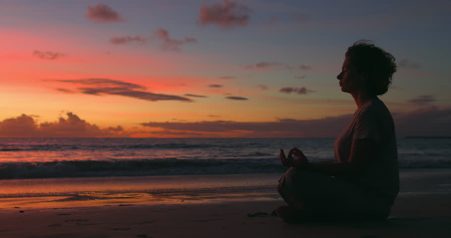 Silhouette of woman in lotus pose sitting on seashore at pink-orange sunset. Curly woman meditates, being in state of relaxed concentration and giving freedom to thoughts, feelings. Meditation by sea Royalty-Free Stock Footage #1100250661