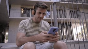 Video of a happy male student using a cell and laughing sitting on the stairs of the university campus. Smiling young boy listening to music and texting and checking social networks on mobile. 