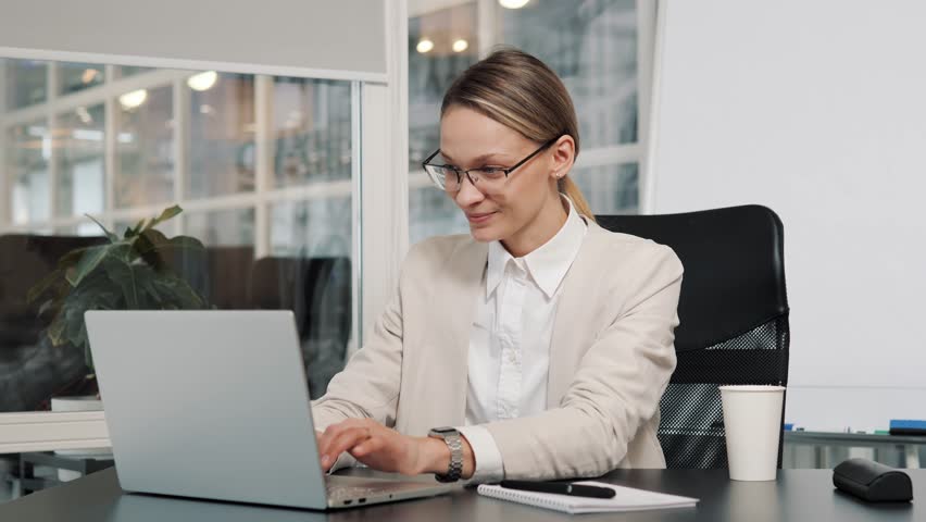 Young business woman manager in glasses and light formal suit sits at table with laptop working from office. Female CEO gets great online news puts hand on chin, satisfied successful entrepreneur. | Shutterstock HD Video #1100254741