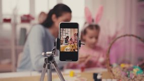 Mother and little daughter decorating Easter eggs, recording video on phone