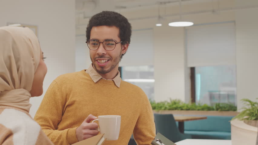 Waist up shot of group of young cheerful Muslim colleagues chatting during coffee break in modern coworking space