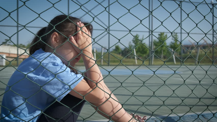 Loneliness of a child. A little girl covering her face with her hands is crying on the sports ground behind the net. The problem of assimilation of refugees. Royalty-Free Stock Footage #1100254997