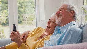 Retired senior couple sitting on sofa in lounge at home together holding remote control watching off camera TV and laughing - shot in slow motion - Powered by Shutterstock - Get 15% off with code: PIKWIZARD15