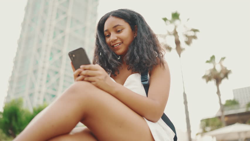 Pre-teenage girl uses mobile phone while sitting on the waterfront. Teenage girl making video call on smartphone | Shutterstock HD Video #1100256045