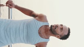 VERTICAL VIDEO: Young bearded male fit athlete standing on the embankment against the sea background, looking at the camera
