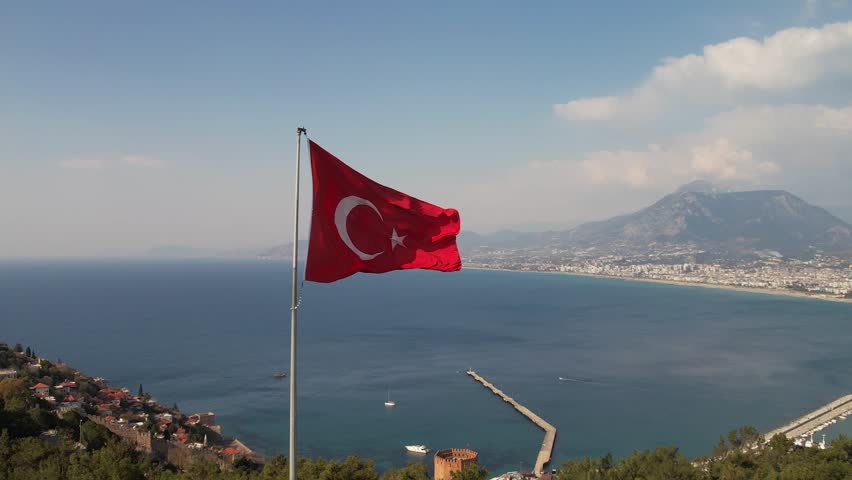 A large flag of Turkey flutters on a flagpole high above the ground against the backdrop of the Alanya fortress and the sea Royalty-Free Stock Footage #1100258421
