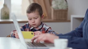 Little Boy Sitting At Table With Phone Near His Mom Who Is Working on Laptop at Home. Kid Plays Mobile Games, Watches Videos and Social Media